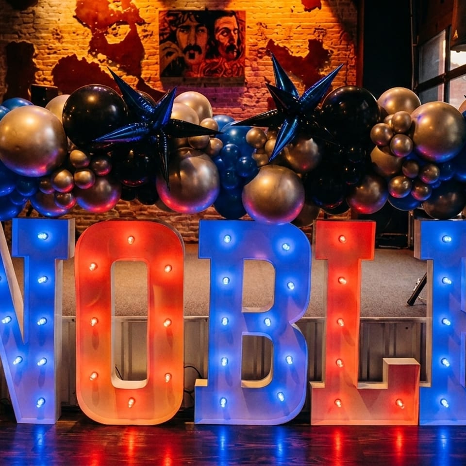 Illuminated red and blue “NOBLE” letters beneath a festive black, gold, and blue balloon arrangement in a brick-walled venue.