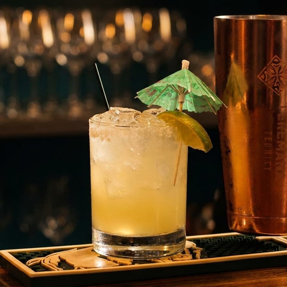 Cocktail with ice, lime wedge, and paper umbrella beside a copper shaker on a dimly lit bar.