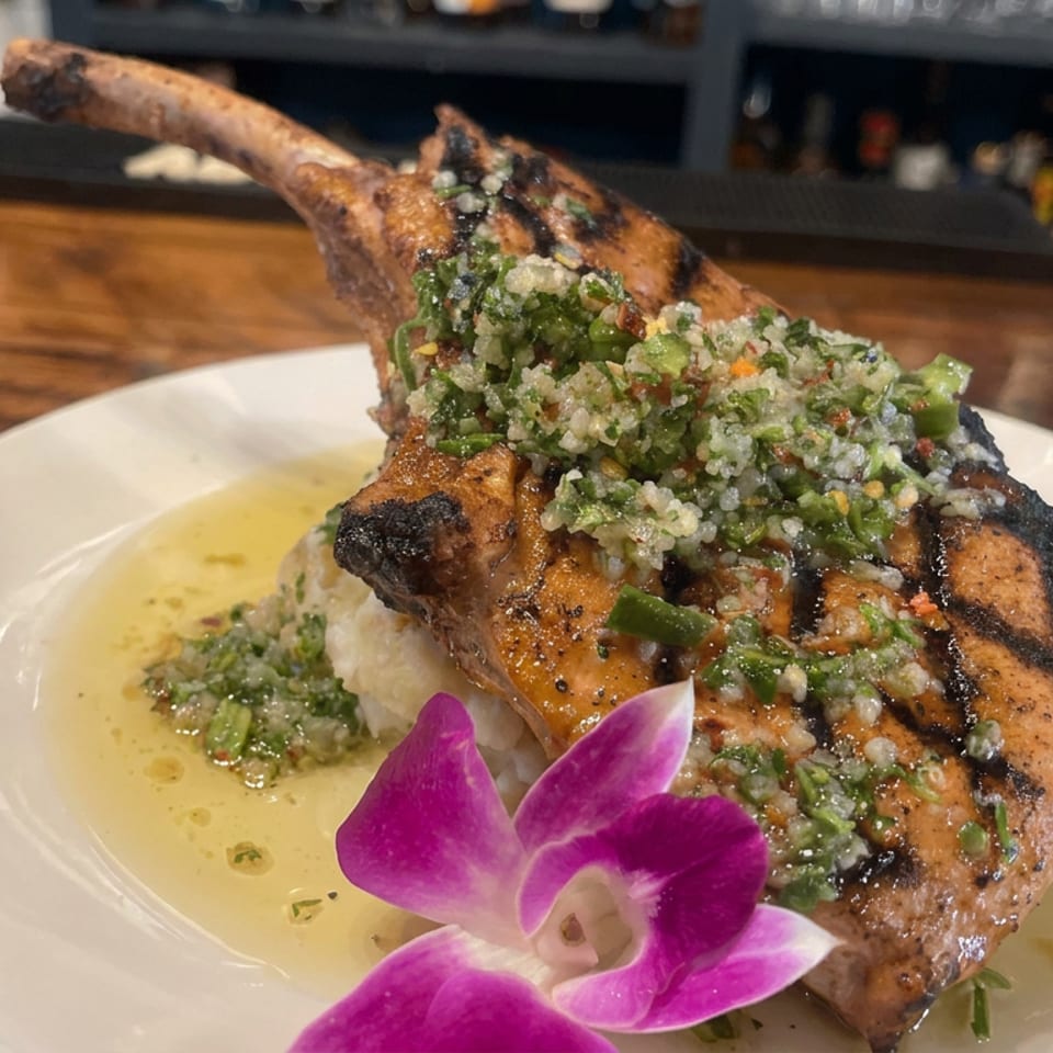 Grilled lamb chop topped with chimichurri, served with rice, asparagus, herb sauce, and an orchid on a white plate.