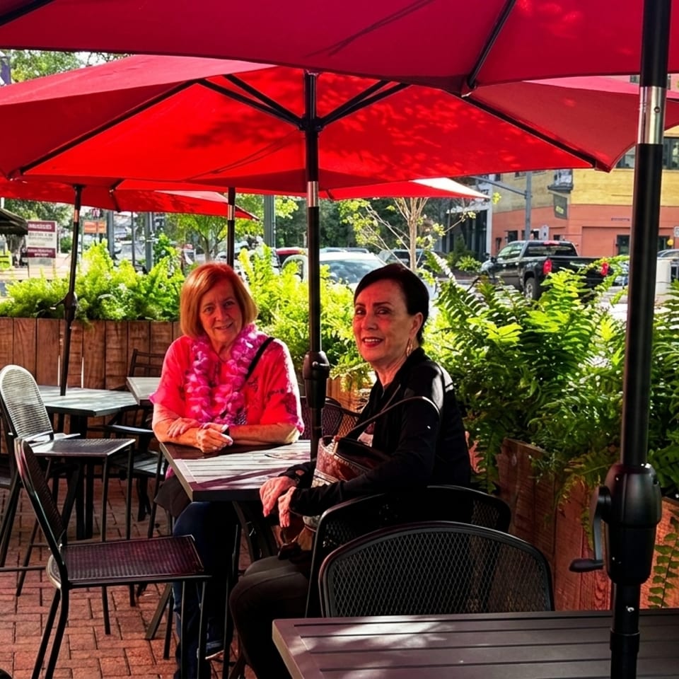 Two women sit at a restaurant’s sidewalk patio table beneath red umbrellas, surrounded by plants and outdoor dining furniture.