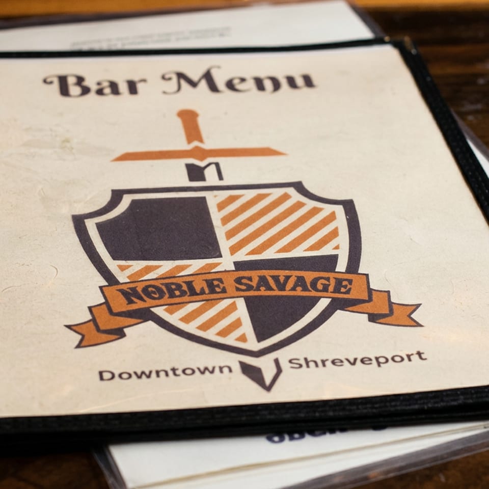 Bar menu featuring the Noble Savage Downtown Shreveport logo on a wooden restaurant table.