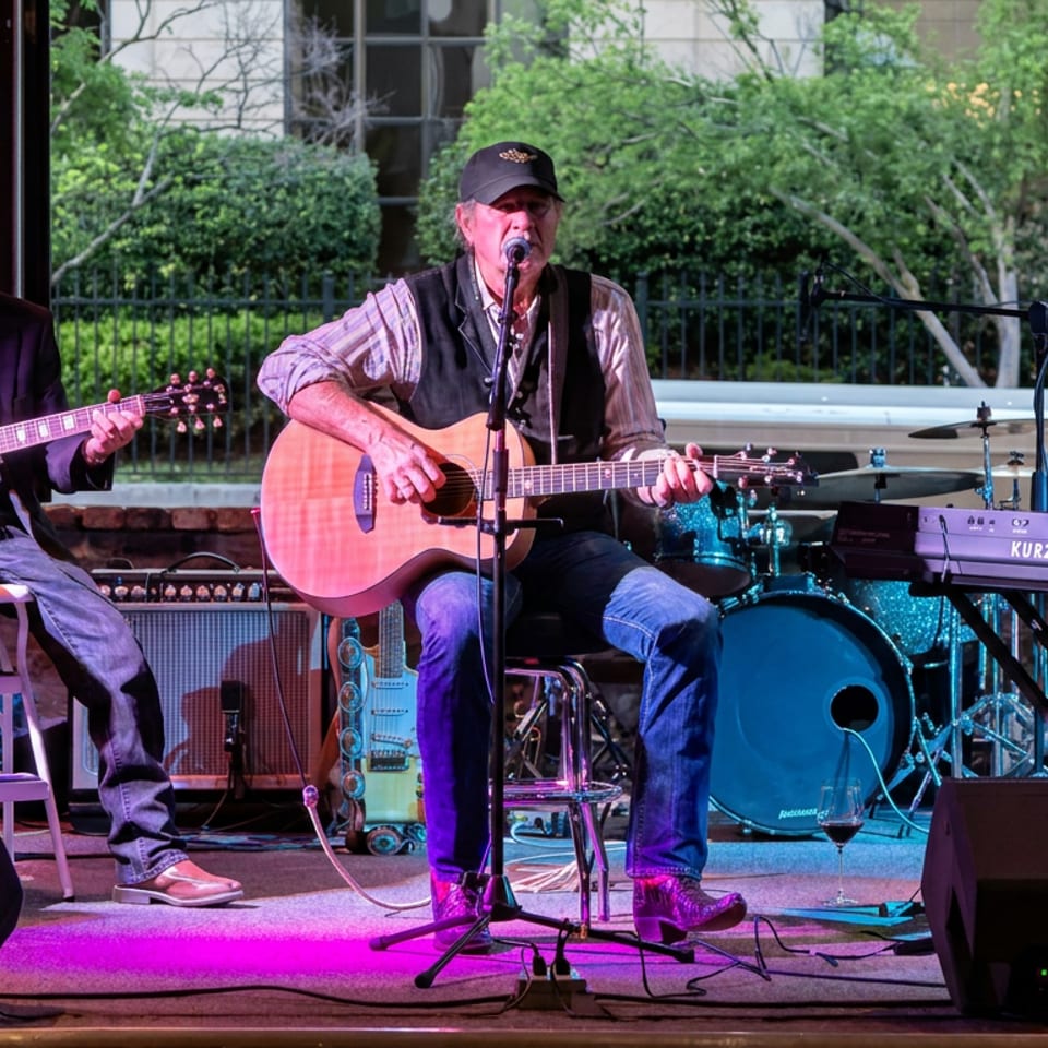 Three musicians perform onstage, playing acoustic and electric guitars, keyboard, and drums in colorful lighting.