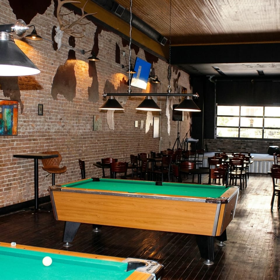 Rustic restaurant bar and dining space featuring two green-felt pool tables, exposed brick walls, pendant lighting, artwork, and wooden tables.