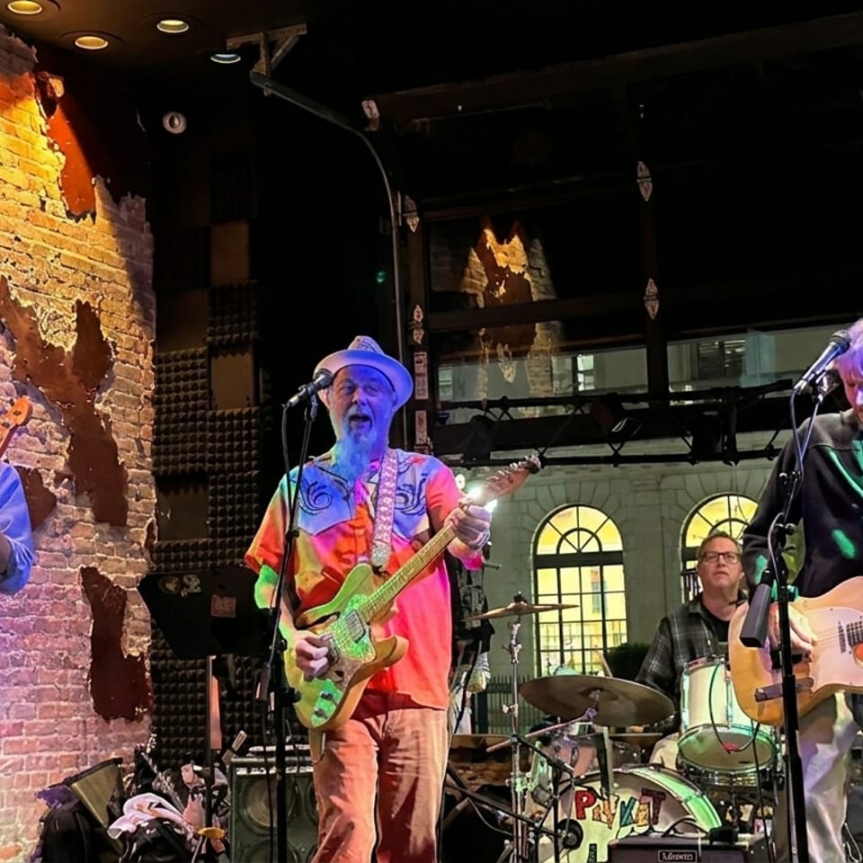 Four musicians perform on a dimly lit restaurant stage, playing guitars and drums beside exposed brick walls.