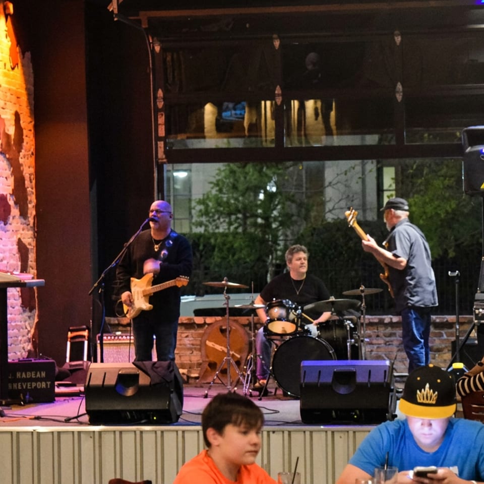 Live band performs on an indoor restaurant stage, featuring guitarists and a drummer, with diners watching.