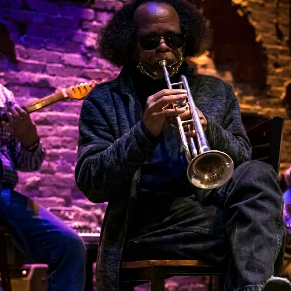 Three musicians perform jazz in a dimly lit brick-walled venue, featuring a trumpet player, guitarist, and bassist under purple lighting.